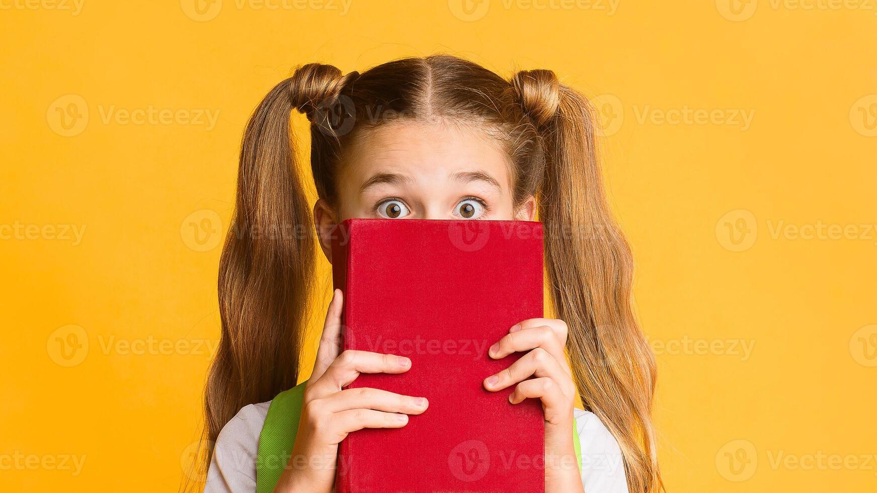 Back To School Anxiety. Scared Elementary Student Girl Hiding Behind Book On Yellow Studio Background. Free Space photo