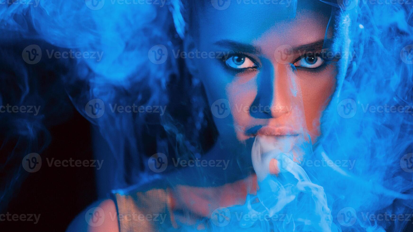 A woman with striking eyes is enveloped in blue smoke while she gazes intensely in a dark room. The blue lighting enhances the dramatic and mysterious atmosphere of the setting. photo