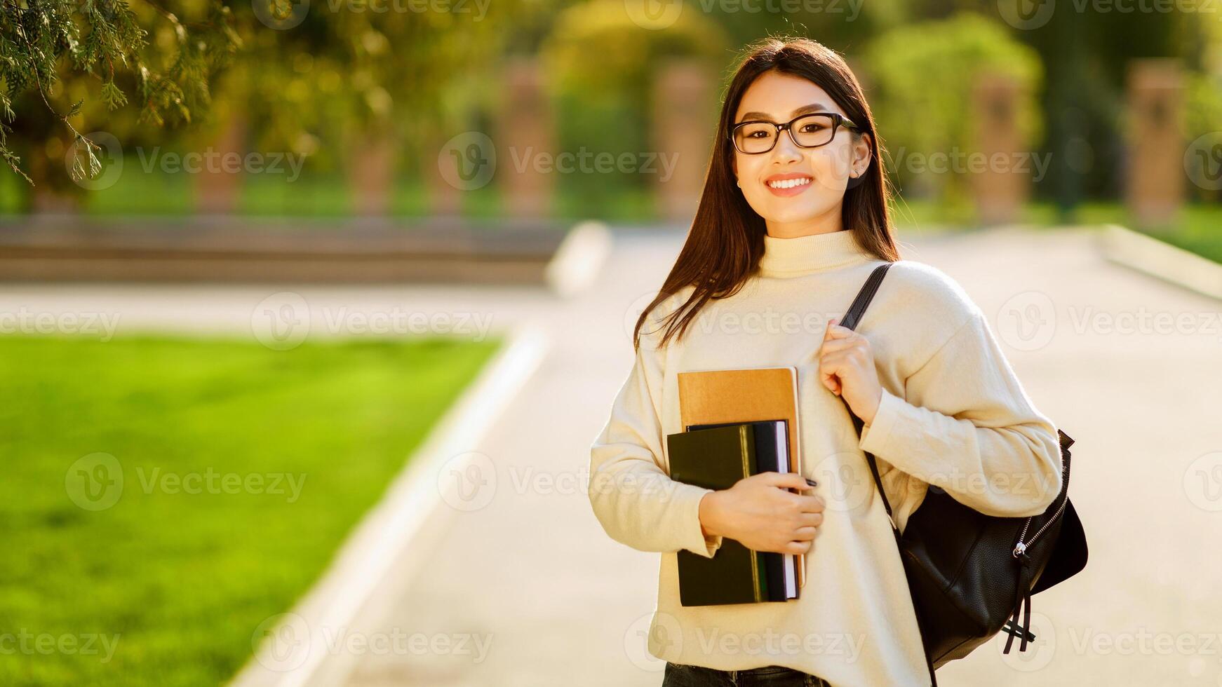 Asian young woman with glasses is walking on a college campus during the morning, holding books and a backpack. She is wearing a light-colored sweater and smiling, copy space photo
