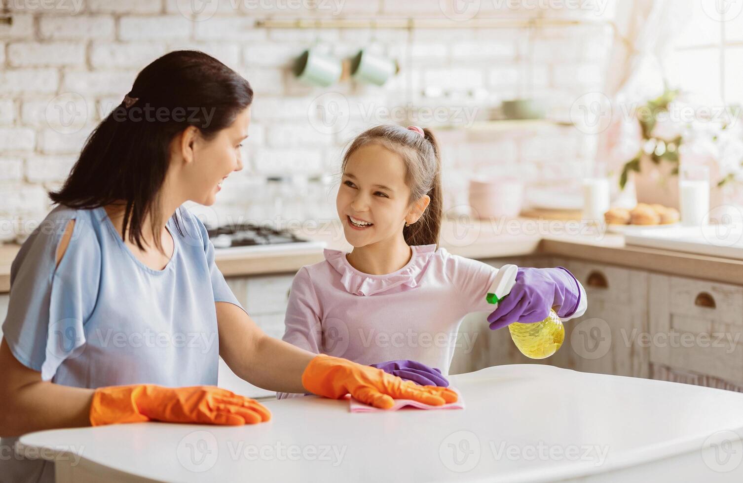 A mother and her daughter are cleaning a kitchen table together. The mother is wearing orange gloves and wiping the table with a cloth. The daughter is wearing purple gloves and spraying the table photo