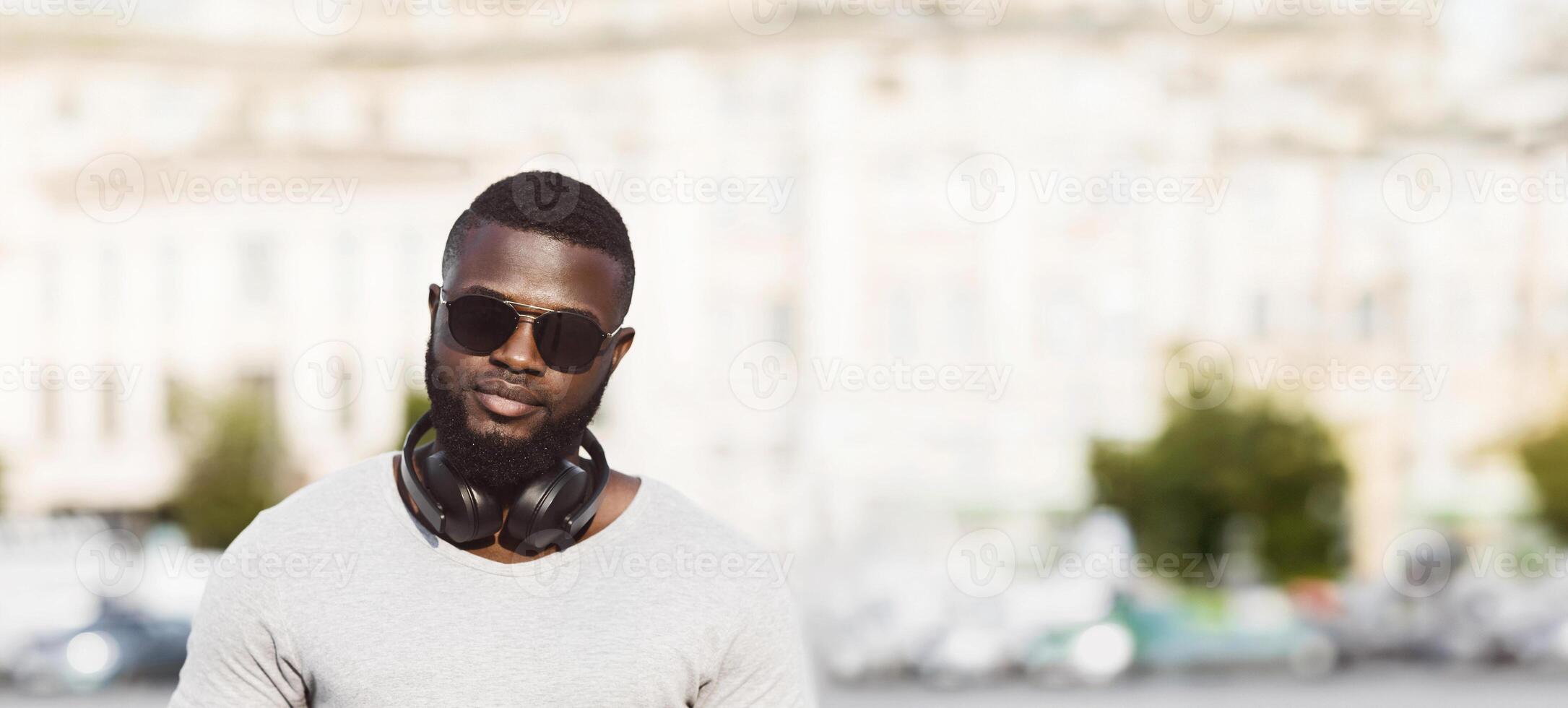 A man with a beard and sunglasses is wearing headphones around his neck. He is looking directly at the camera while standing in front of a blurred city background. photo