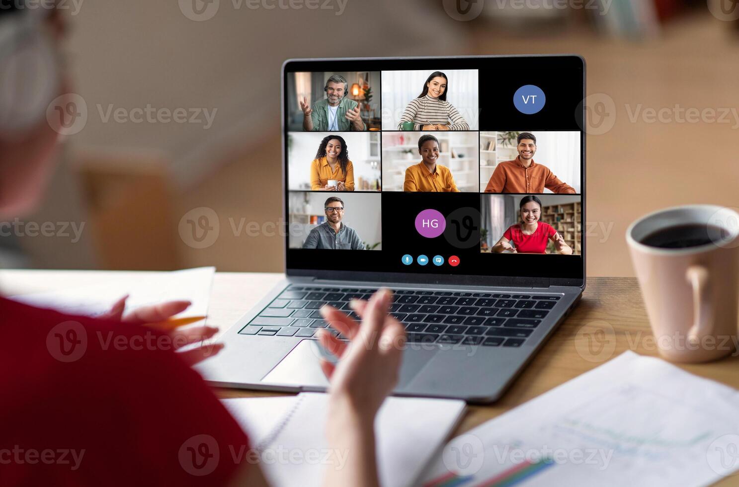 A woman sits at her desk, participating in a conference call on her laptop. The screen displays multiple participants, with their names and initials visible photo