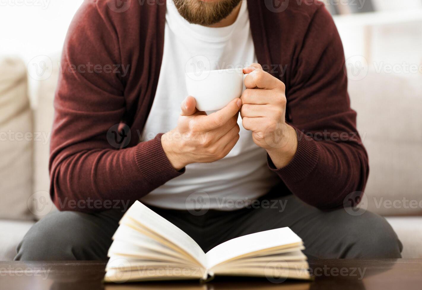 A man is seated on a couch, holding a cup in one hand and engrossed in reading a book. He appears relaxed and focused on the text, with a comfortable posture, cropped photo