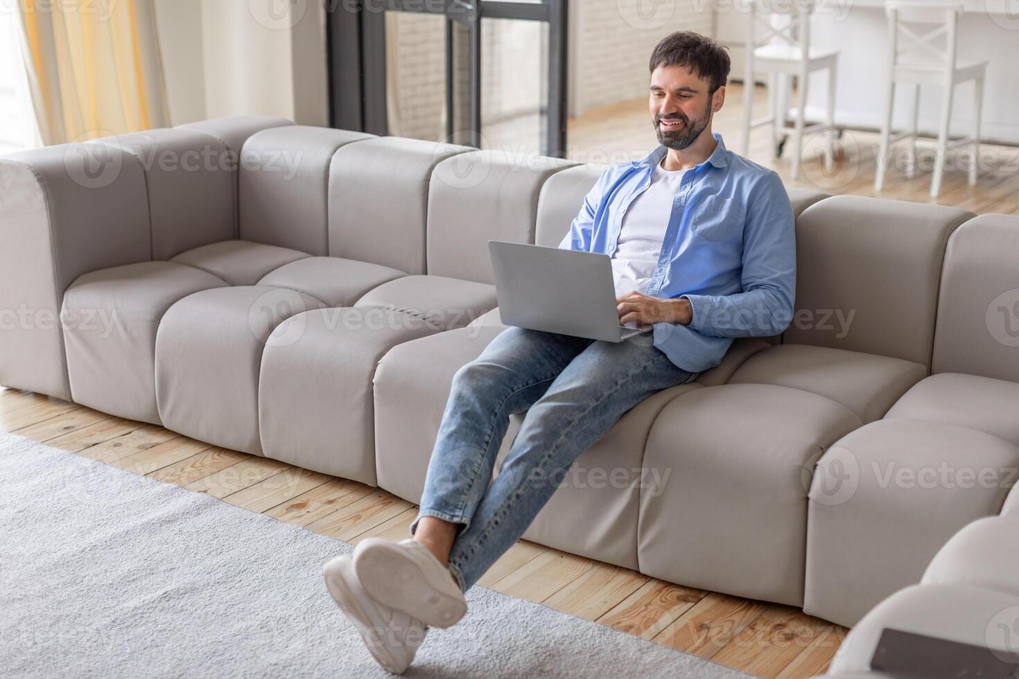 A man sits on a grey modular sofa in a modern living room, using a laptop computer. He is smiling and looks relaxed, possibly enjoying a break from work, high angle view photo