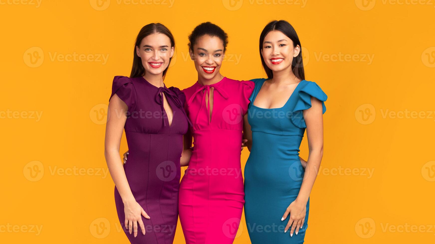It Girls. Three Diverse Young Ladies In Cocktail Dresses Smiling At Camera And Hugging Standing On Yellow Background. Studio Shot photo