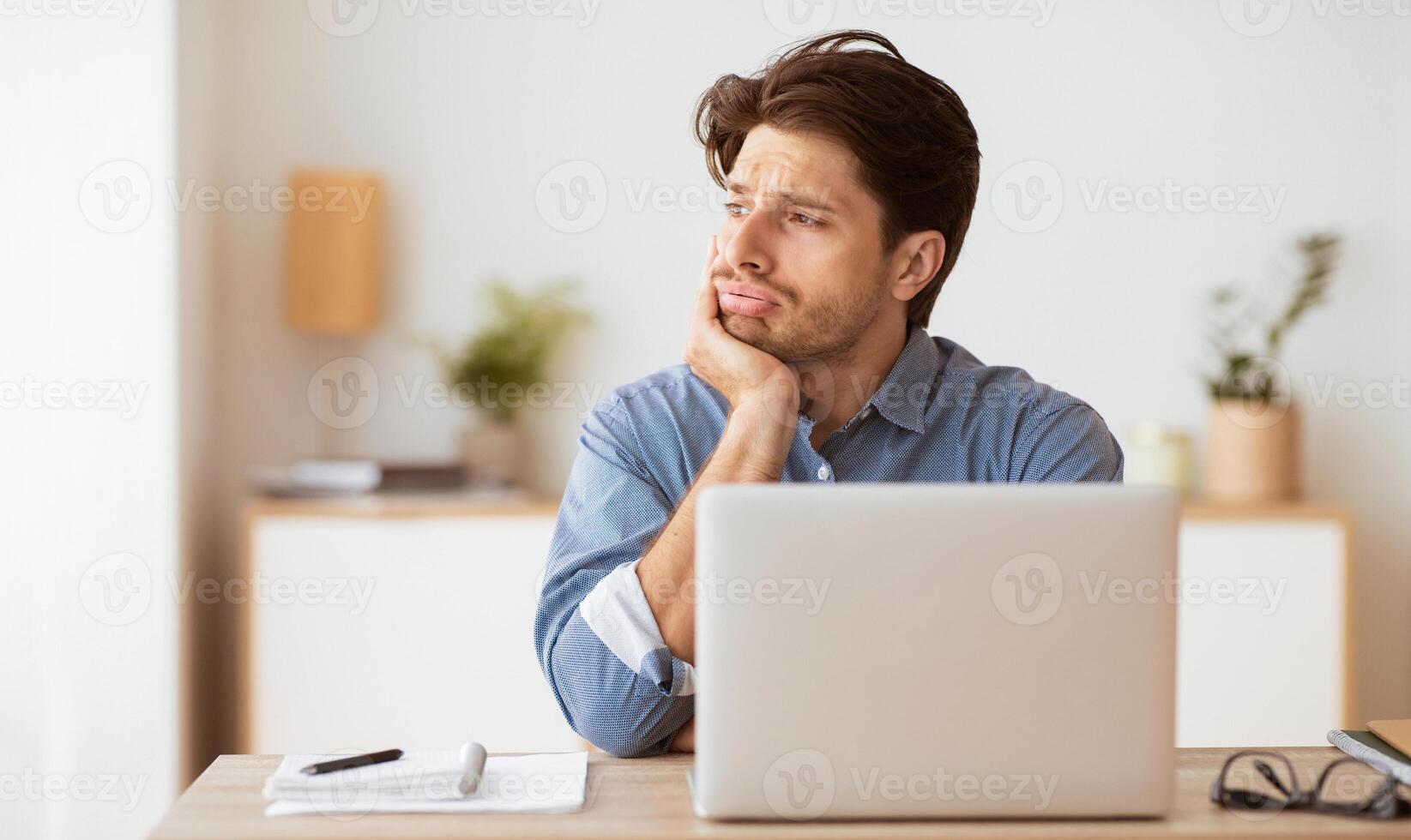 A pensive young man gazes out the window at his desk, chin on hand, with a laptop in front of him and a notepad and pen nearby. photo