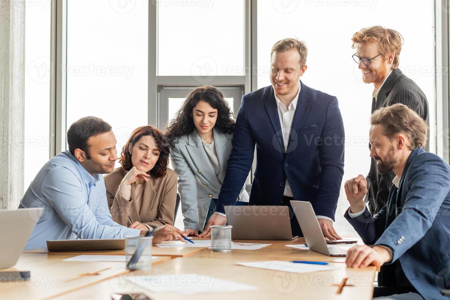 A group of diverse business professionals are gathered around a table in a modern office, working together on a project. They are looking at a laptop screen and discussing ideas photo