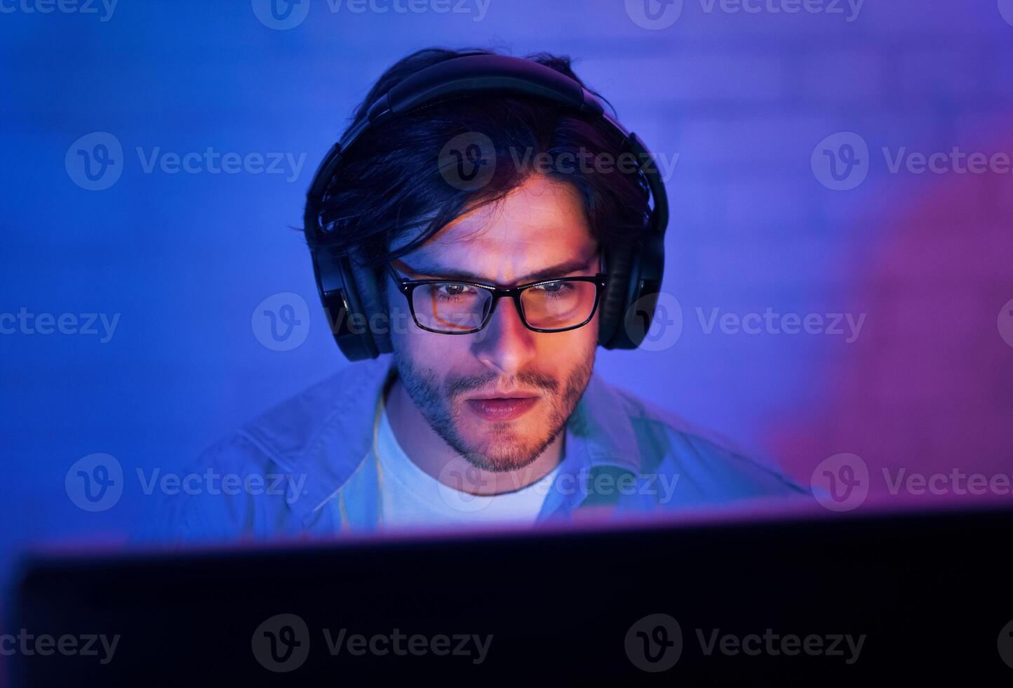 A young man wearing glasses and headphones sits in front of a computer screen in a dimly lit room. He appears to be playing a game, looking intently at the screen. photo