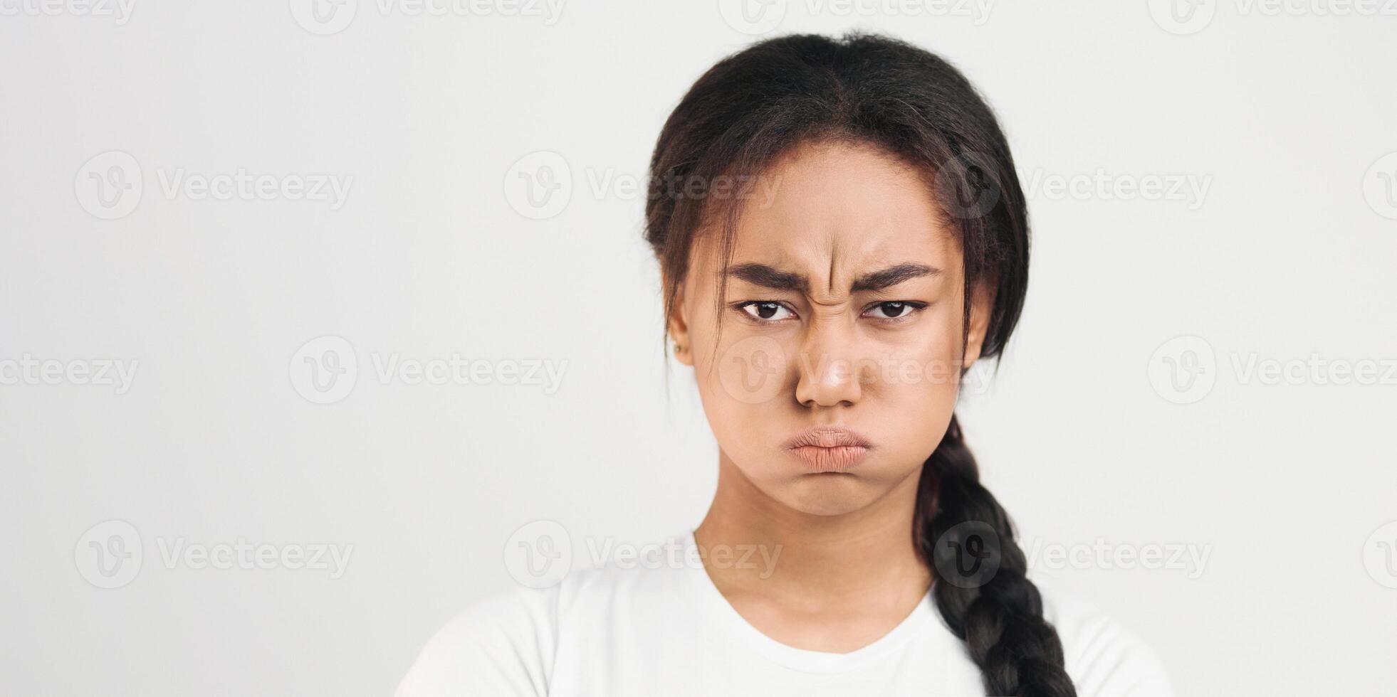African American young girl with a braided ponytail is displaying an expression of anger on her face, with narrowed eyes and a furrowed brow. She appears to be in a state of frustration or irritation. photo