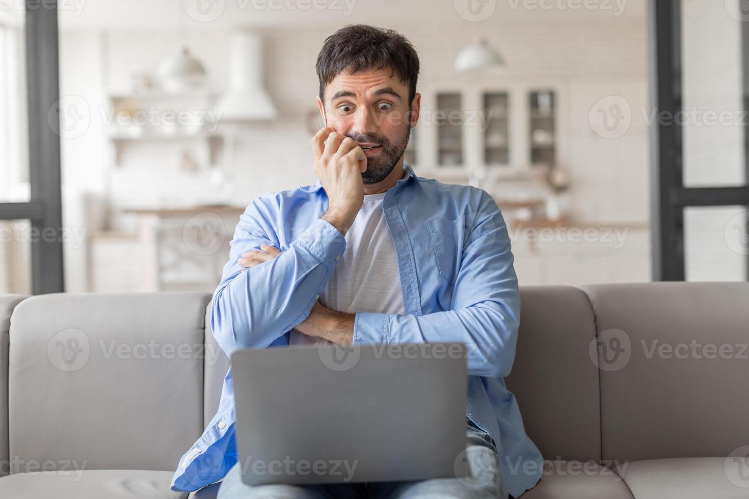 Man sitting on a couch in a home setting. He is looking at his laptop with a worried expression on his face and has his hands clasped together in front of him. He appears to be stressed or anxious. photo