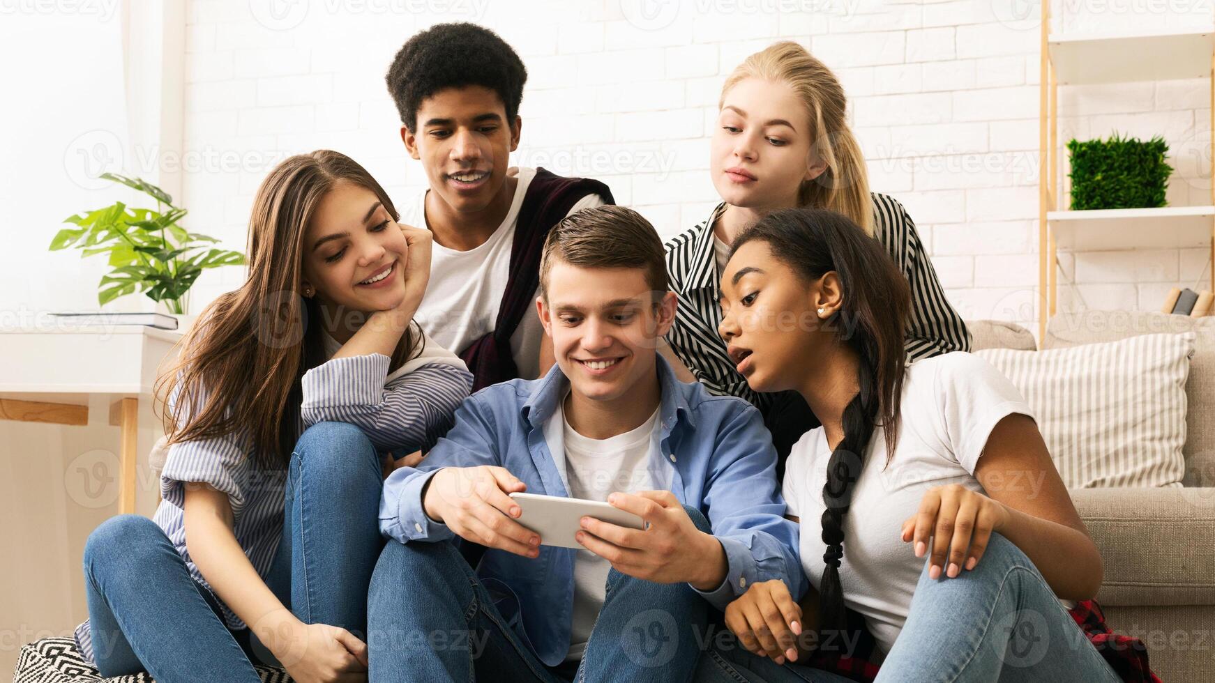 A group of diverse friends sitting closely together on a couch, all engrossed in looking at a cell phone screen. They appear focused and engaged in the content displayed on the device. photo