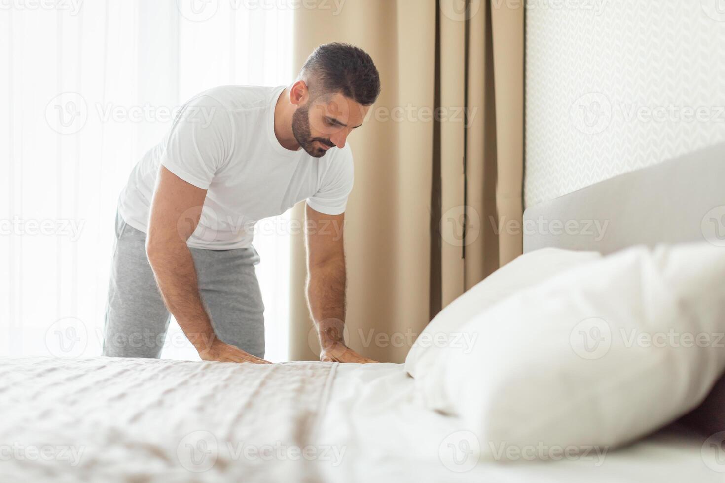 A man in a white t-shirt and gray pants is making his bed in a modern bedroom. He is standing on the side of the bed and has just finished tucking the sheets. White curtains are in the background. photo