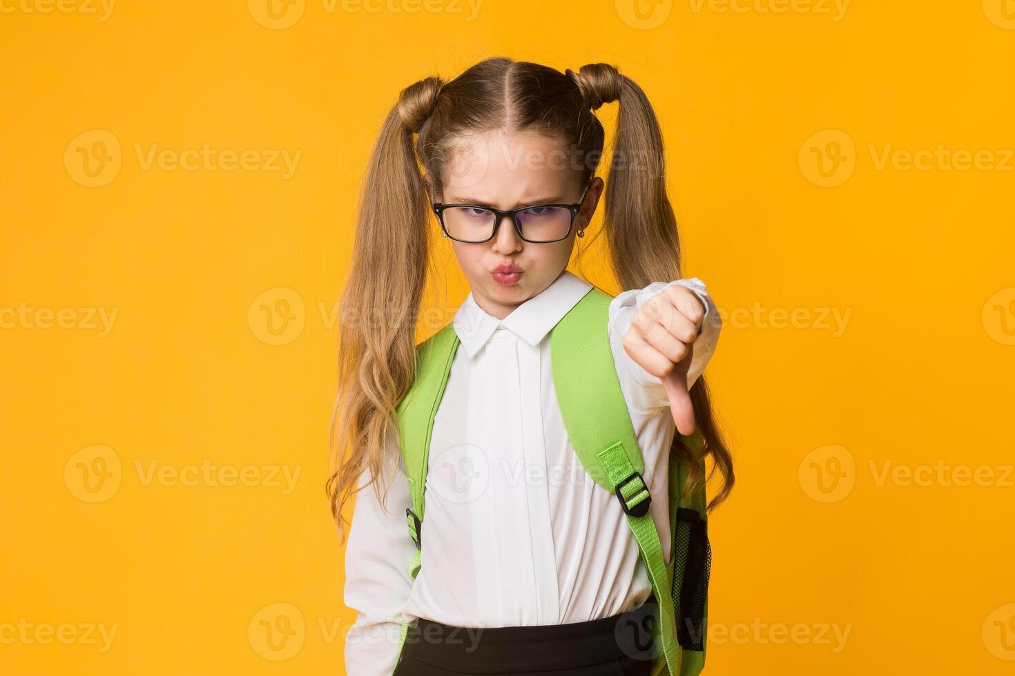 Displeased Schoolgirl Gesturing Thumbs Down In Dislike Disapproving School Posing Over Yellow Background In Studio photo