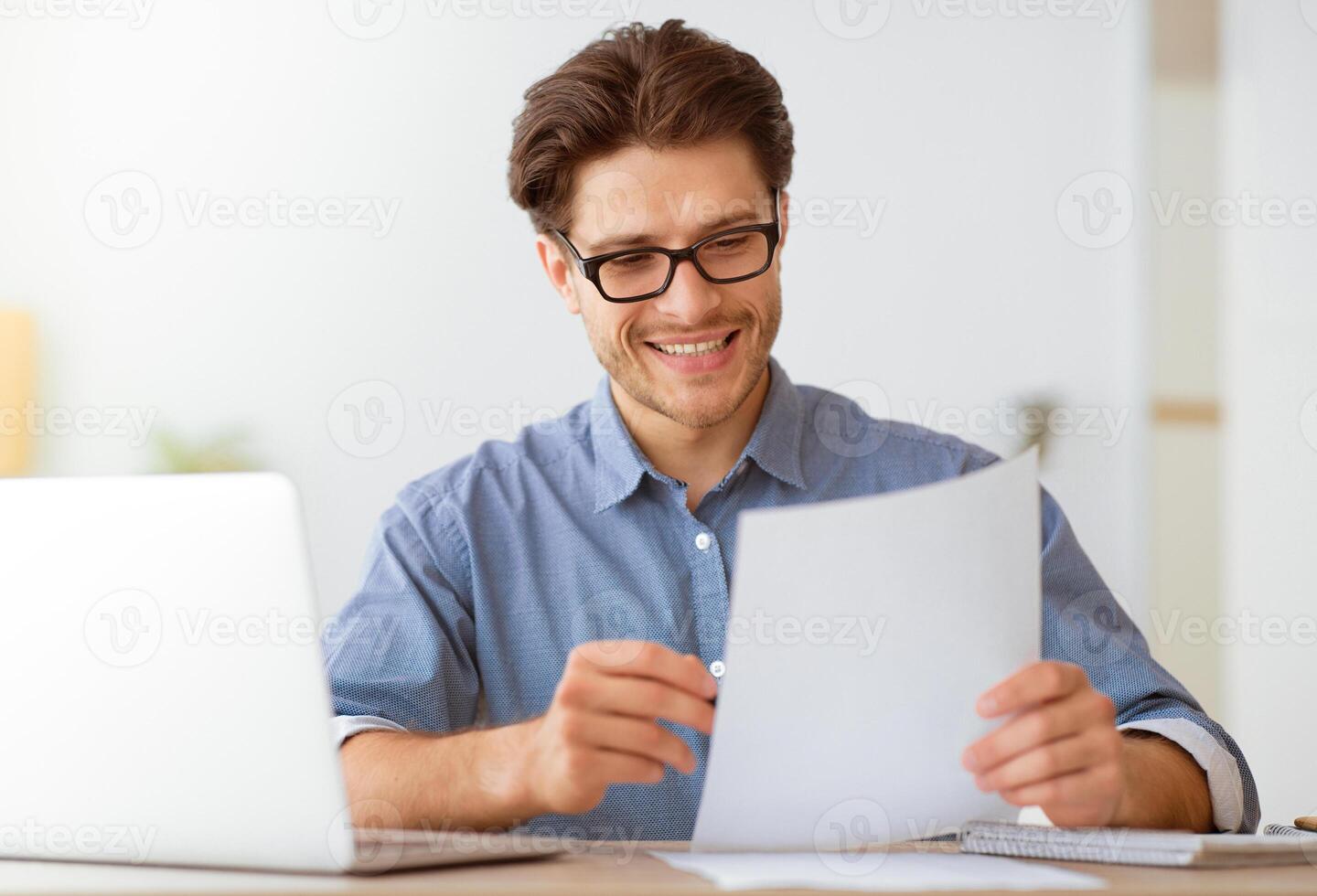 A man smiles as he reads a document while sitting at a desk with a laptop. photo