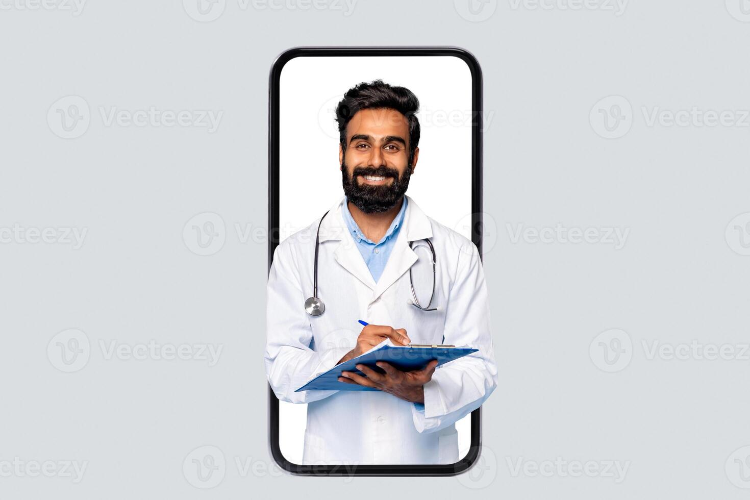 Indian man doctor is seen on the screen of a smartphone, wearing a white coat and stethoscope, smiling, and writing on a blue clipboard with a pen. photo