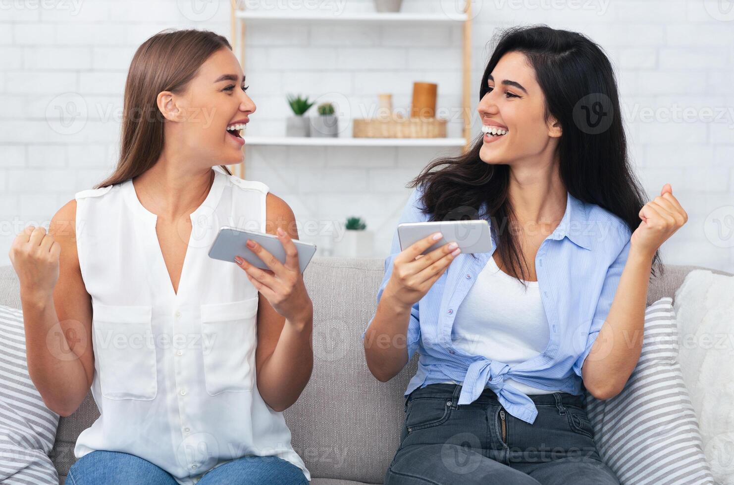 Modern Friendship. Two Girls Playing Mobile Games On Phones Gesturing Yes Celebrating Victory Sitting On Couch Indoor. photo