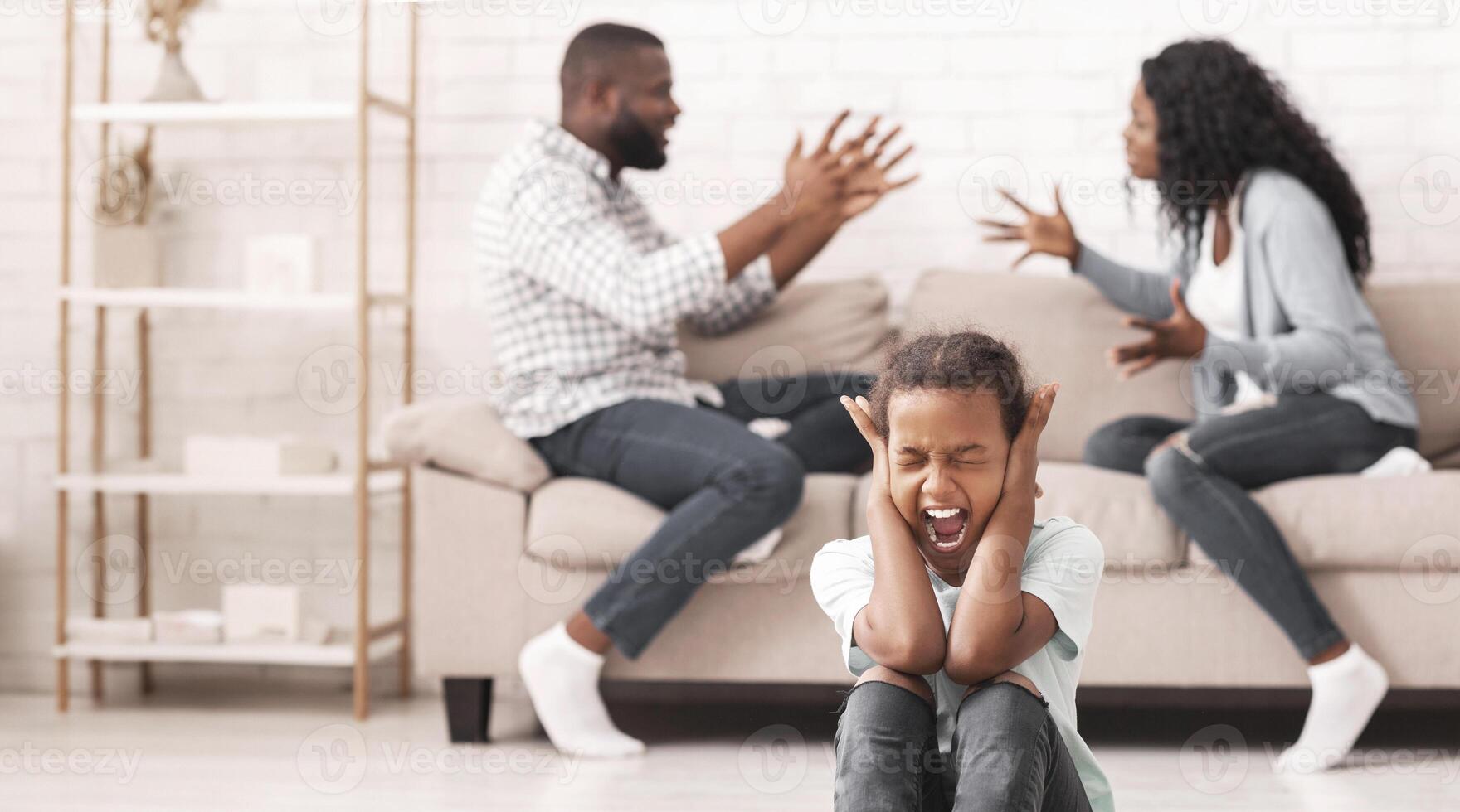Defensive reaction. Upset little black girl sitting on floor, yelling and closing ears not to listen parents arguing, selective focus on child photo