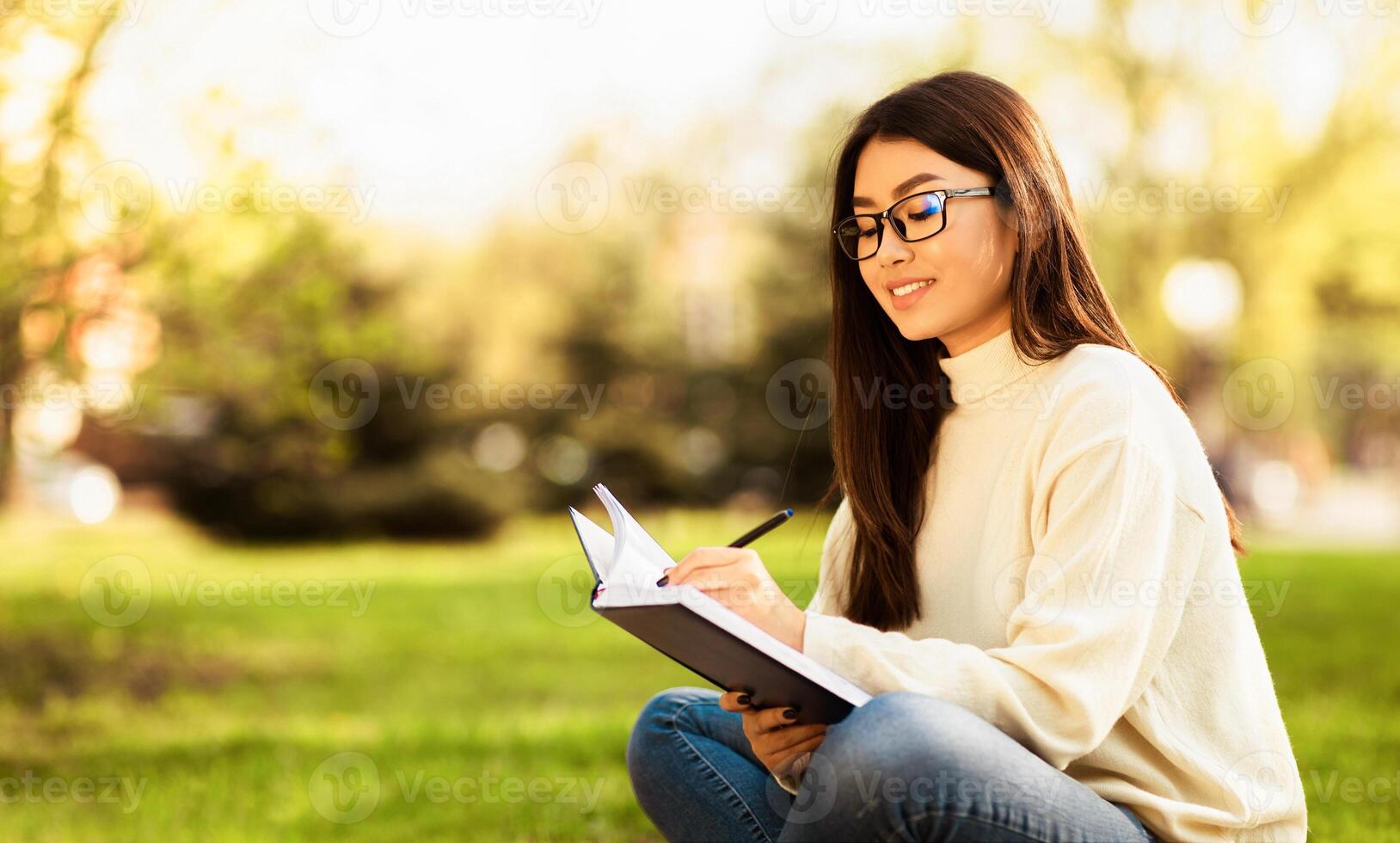 Asian woman is seated on the grass, focused on writing in a notebook. She holds a pen in her hand, with an open notebook on her lap. photo