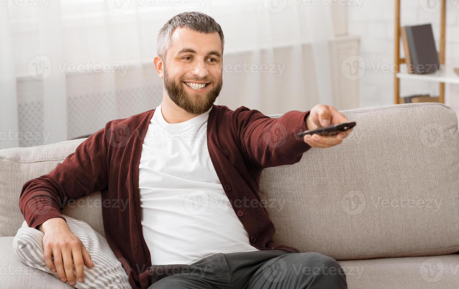 A man is seated on a couch, holding a remote control in his hand. He appears to be focused on the television or other electronic device in front of him. The room is casually furnished photo