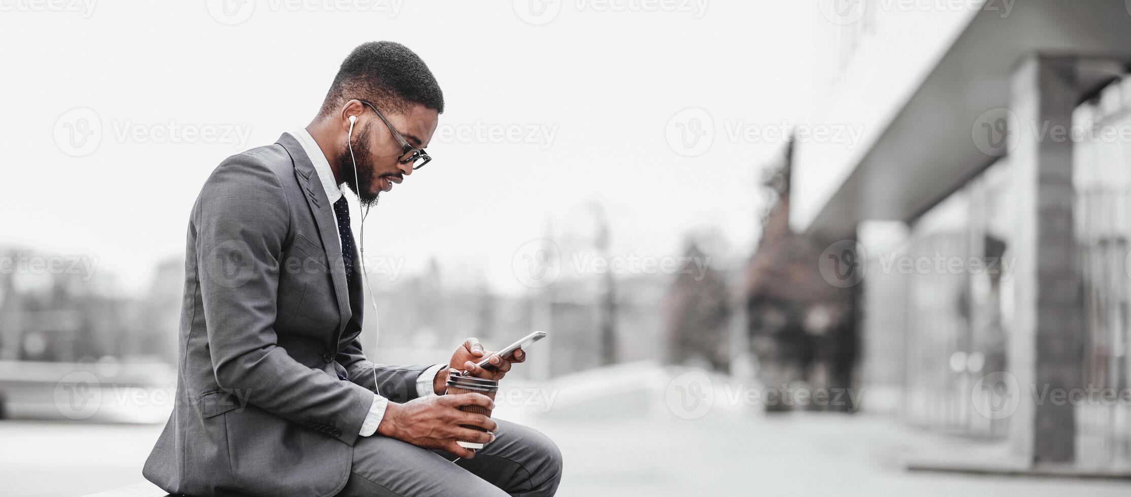 Black businessman with coffee and earphones, using smartphone while sitting outdoors in an urban setting, panorama with copy space photo