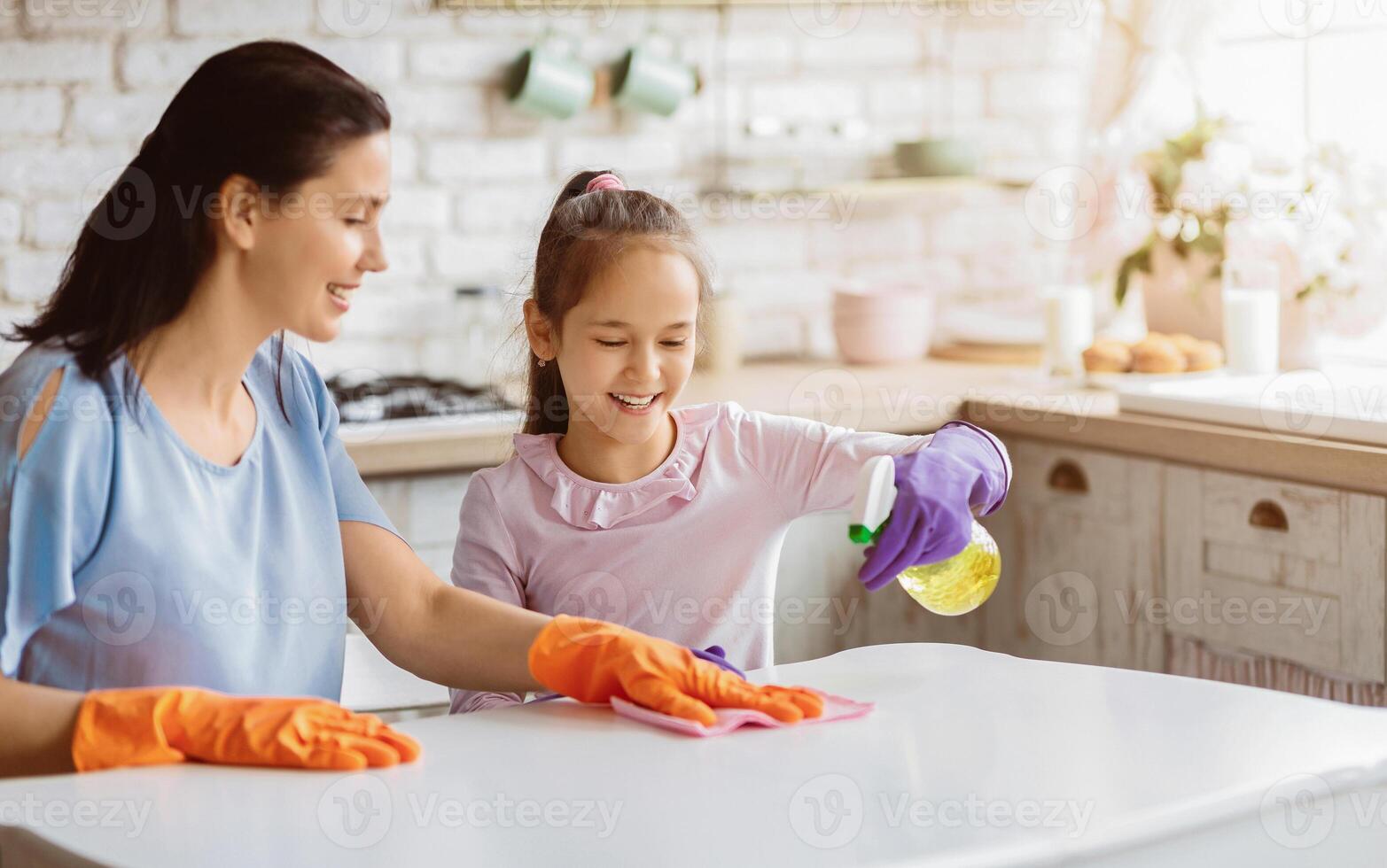 A mother and her young daughter are cleaning their kitchen table together. TThey both have happy expressions on their faces, suggesting that they are enjoying their time together. photo