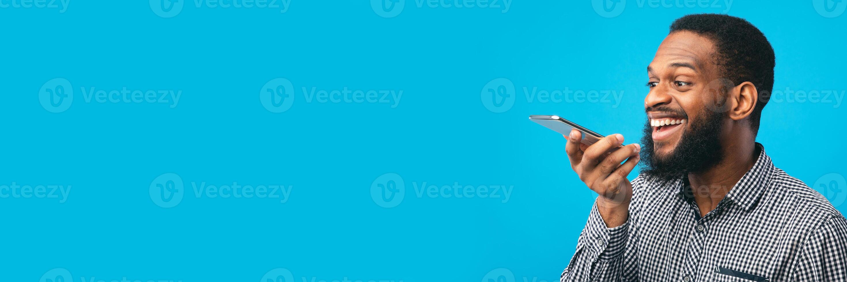 Happy young black man using voice assistant on smartphone, isolated over blue studio wall, copy space, web-banner photo