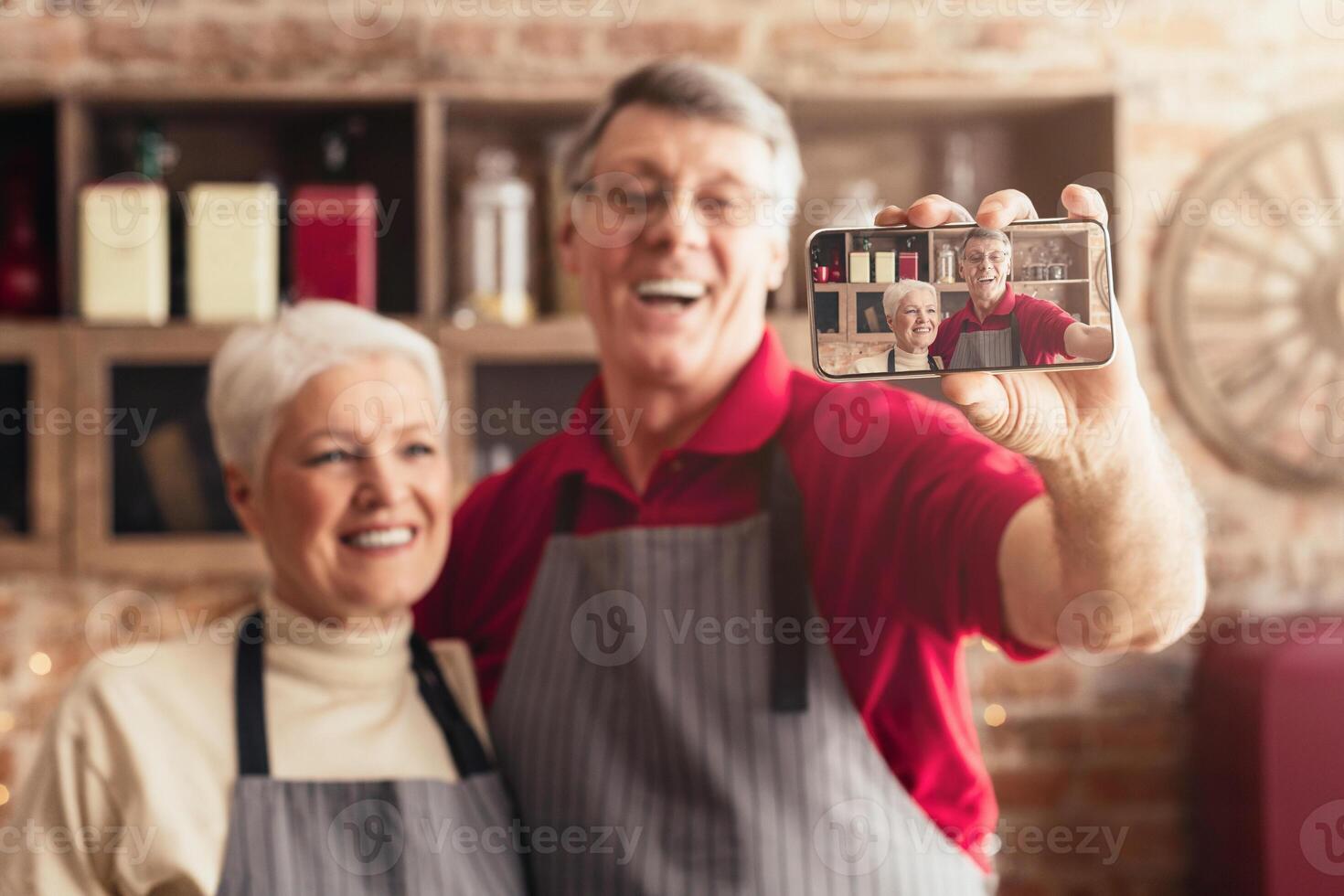An older couple wearing aprons is taking a selfie in their kitchen. They are smiling and appear to be enjoying themselves while preparing a meal together. photo