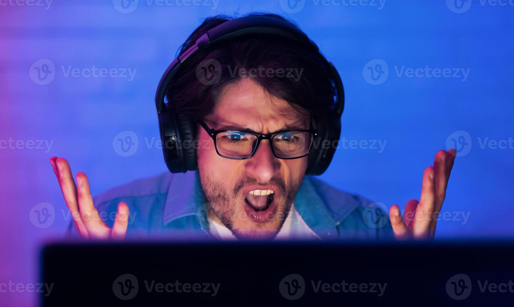 A young man wearing headphones looks frustrated while sitting in front of a computer screen in a room with blue lighting. He is looking directly at the camera with his mouth open in a scream. photo