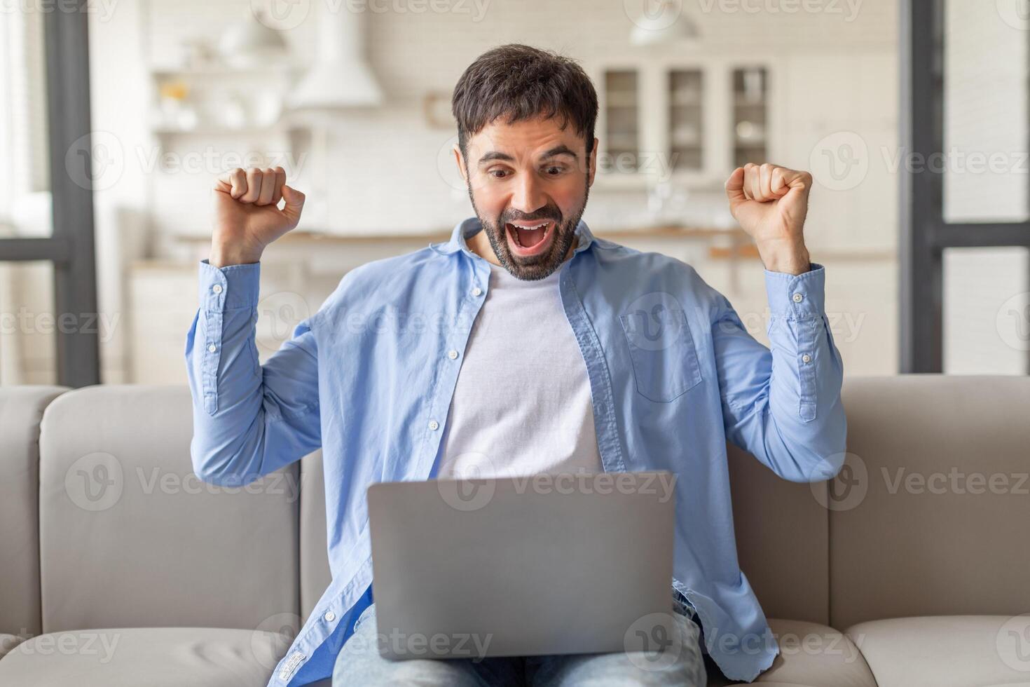 A man sits on a couch, smiling widely and raising both fists in the air, while looking at a laptop screen. He appears to be celebrating a victory or success. photo