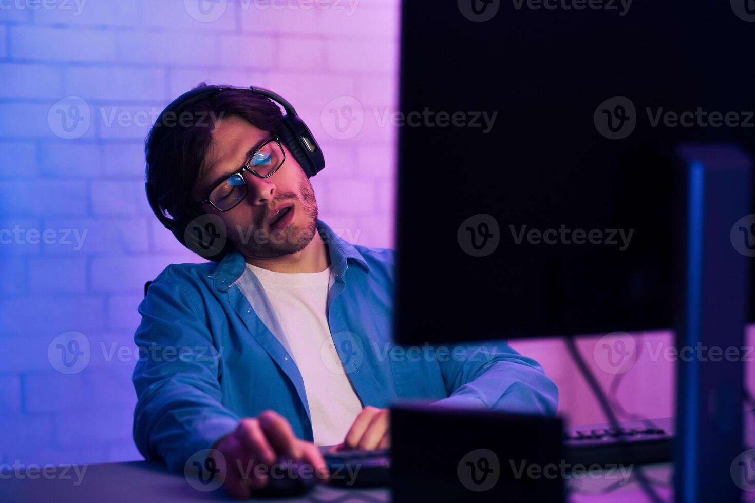 A young man wearing glasses and headphones is asleep at a computer desk. He is sitting in a chair with his head resting on his hand. The desk is lit by a pink and blue light photo
