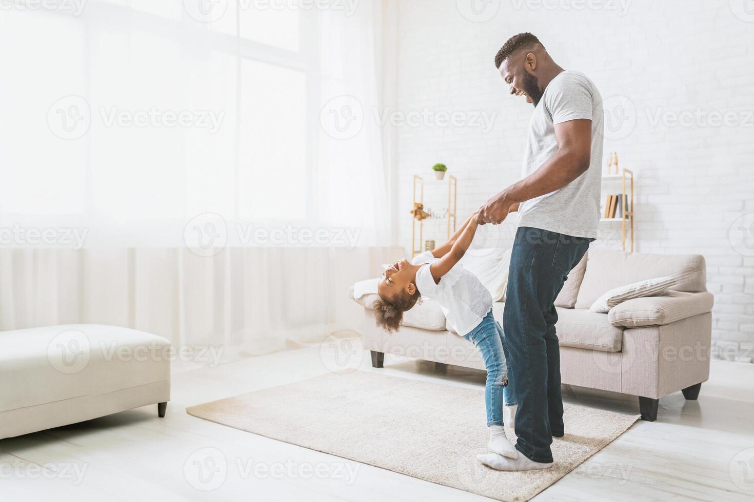 African American happy father spins his daughter around in the living room. The little girl is laughing and has her arms stretched out as her father holds onto her hands, copy space photo