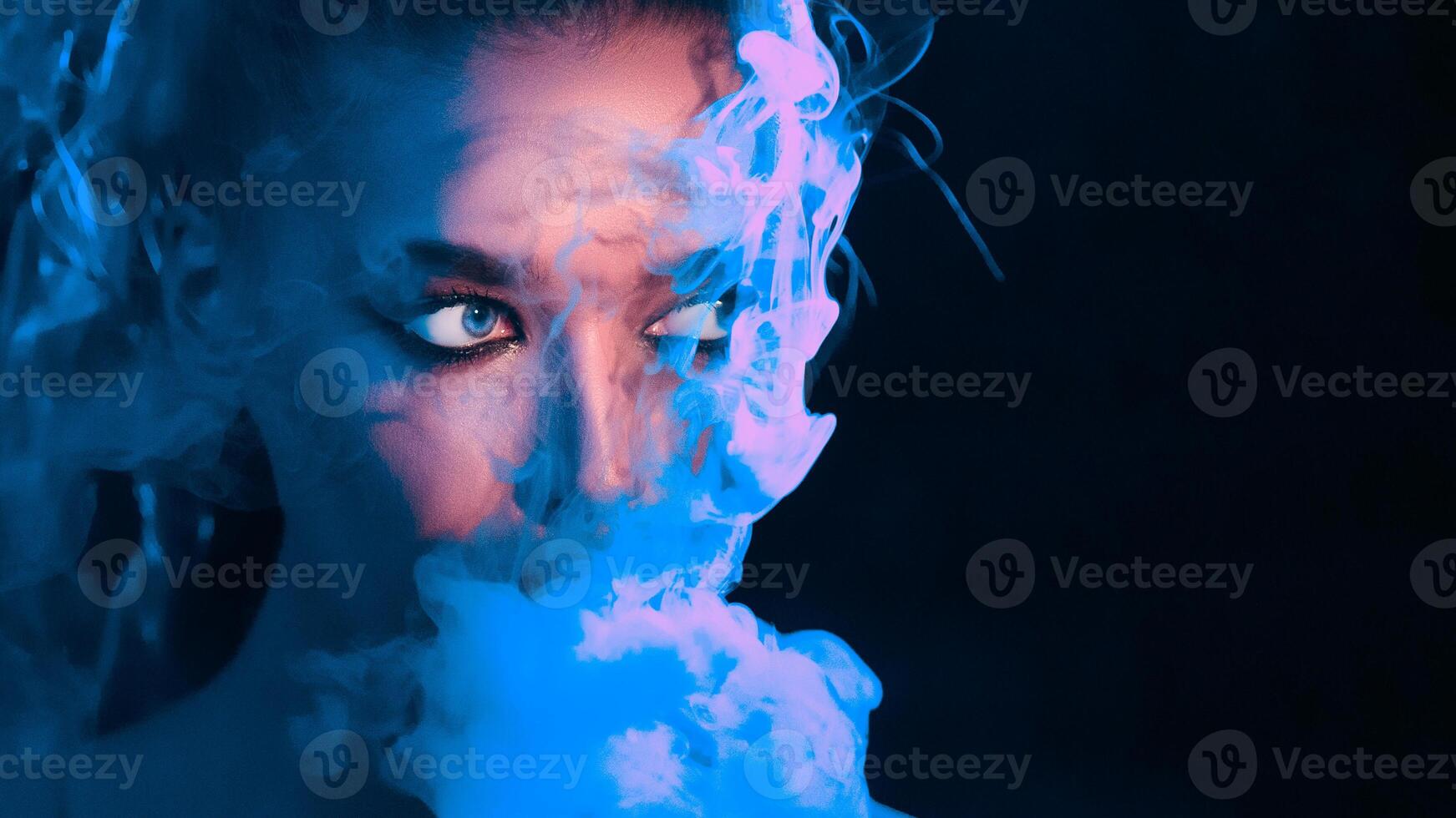 A woman with a captivating, intense gaze peers through swirling blue smoke, creating an air of mystery and intrigue in a dark setting, copy space photo