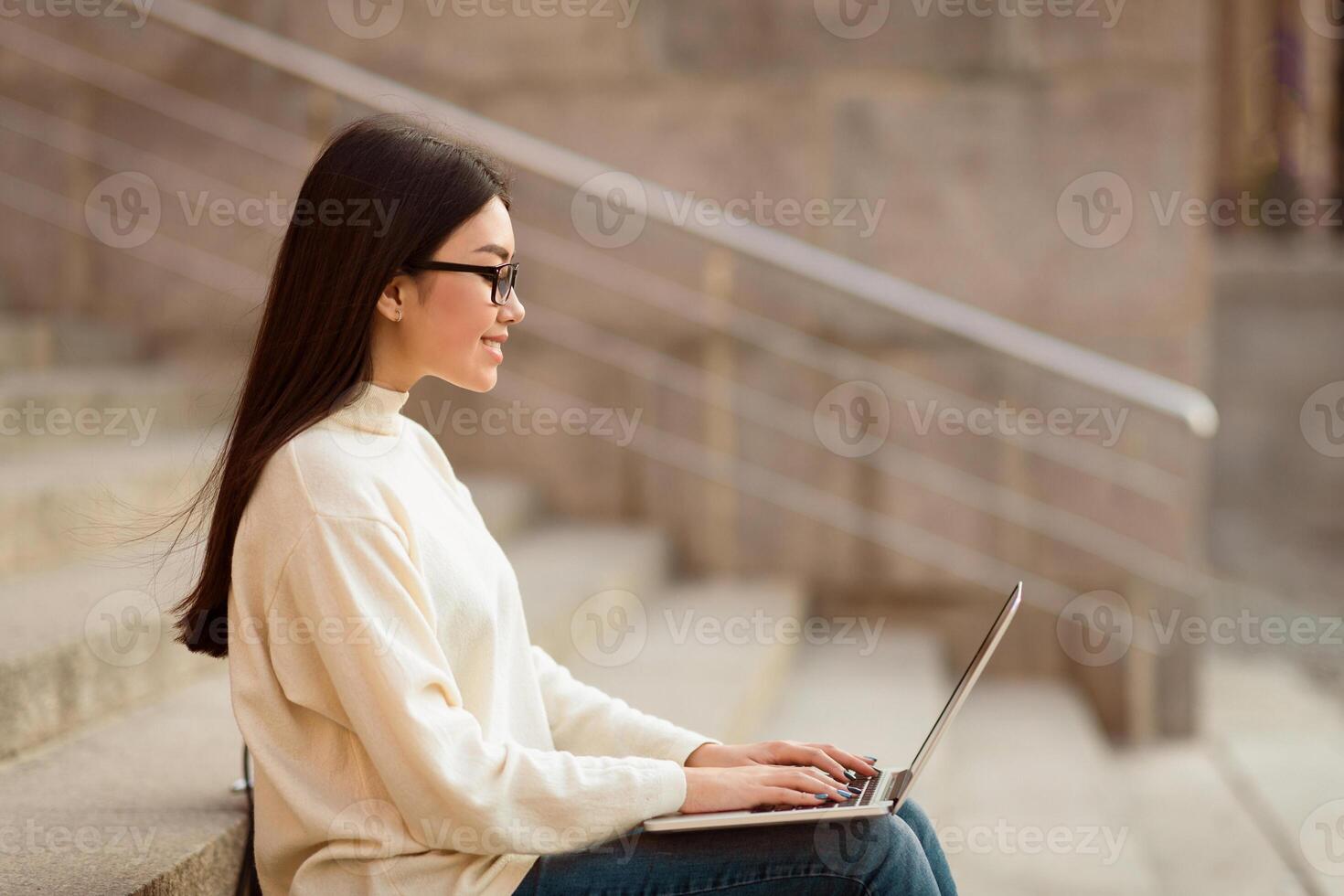 Asian woman is seated on a ledge, focused on her laptop computer. She appears concentrated and engaged in her work or task, with the cityscape visible in the background, blank space photo