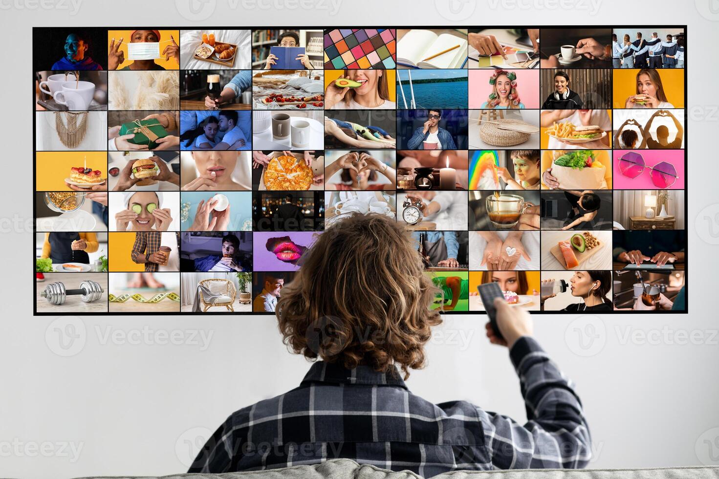 A person sits on a couch facing a television screen, holding a remote in their hand. The television is displaying a grid of colorful images, each showing a different scene, person, or object photo