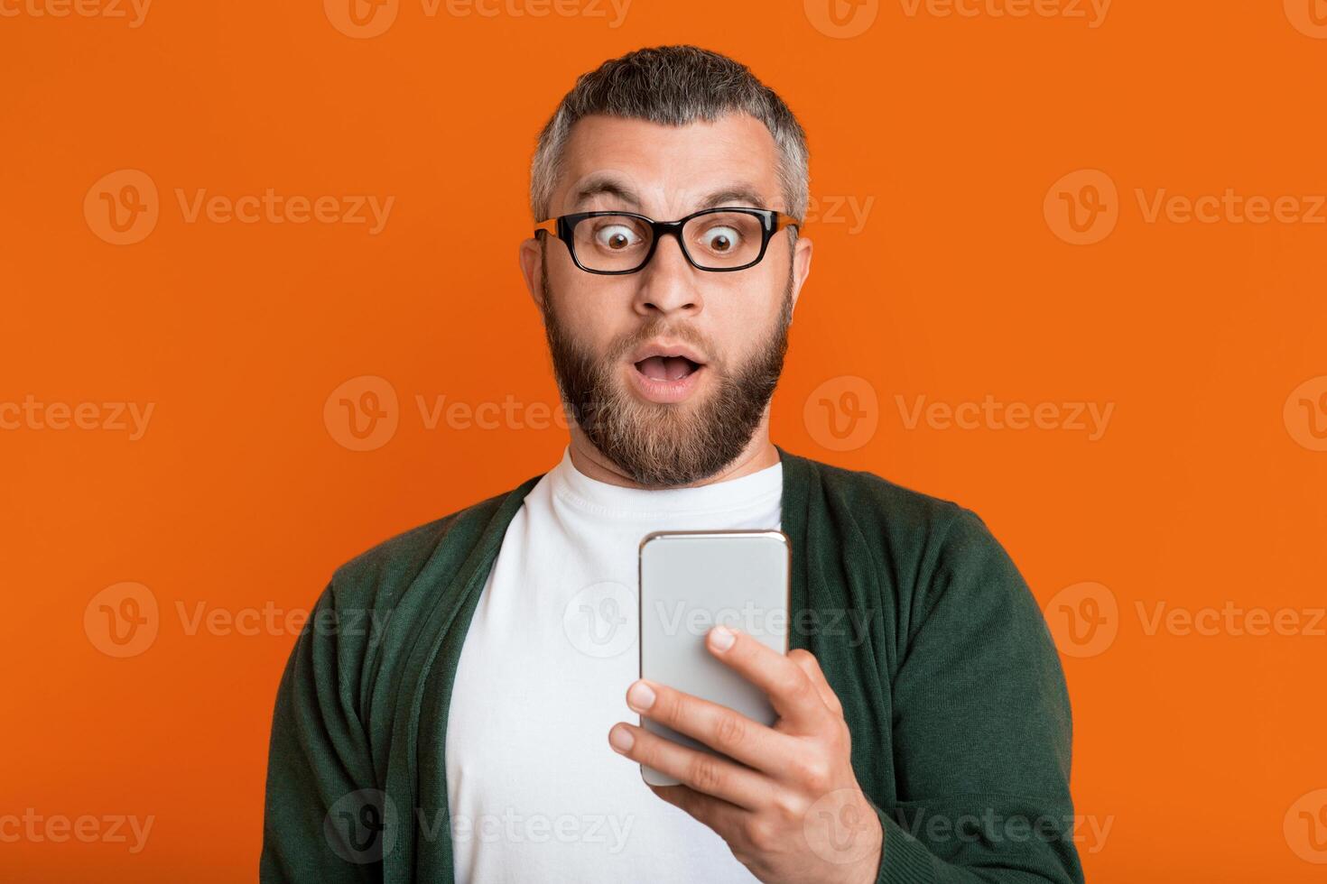 A bearded man wearing glasses is looking at his smartphone with a surprised expression. He stands in front of a bright orange backdrop, wearing a green sweater over a white shirt. photo