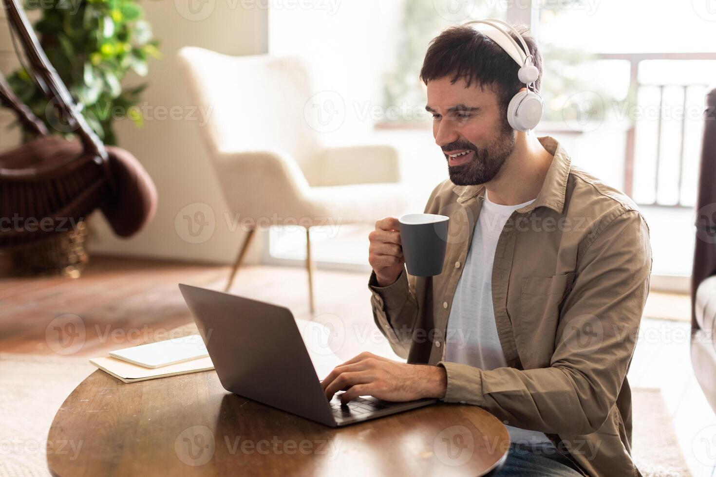 A man is seated at a table, focused on his laptop screen. Next to him is a cup of coffee, steam rising. The mans hands are typing on the keyboard as he works. photo