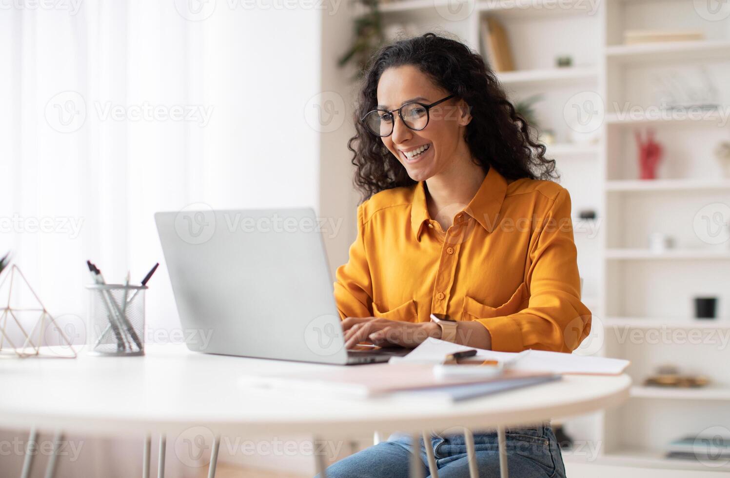 Happy Middle Eastern Businesswoman Using Laptop Typing And Working Online Sitting At Workplace Wearing Eyeglasses. Office Worker Lady Browsing Internet On Computer. Entrepreneurship. Side View photo