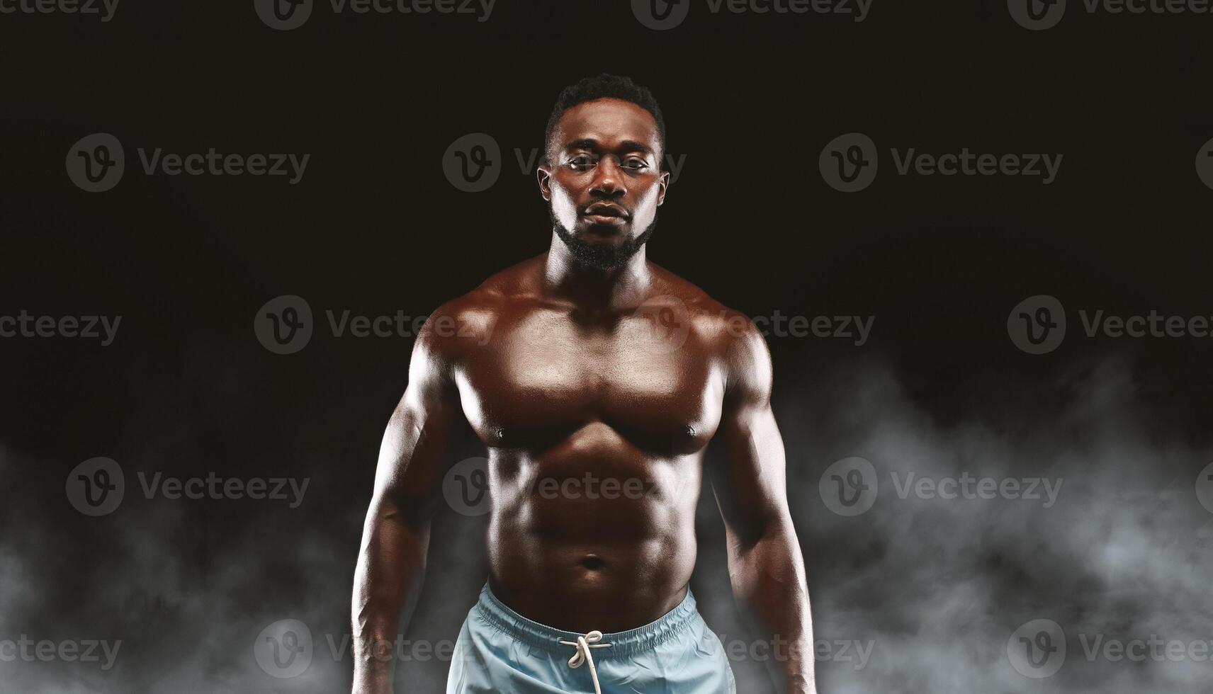 A muscular man poses with his arms at his sides in front of a dark background with smoke. photo