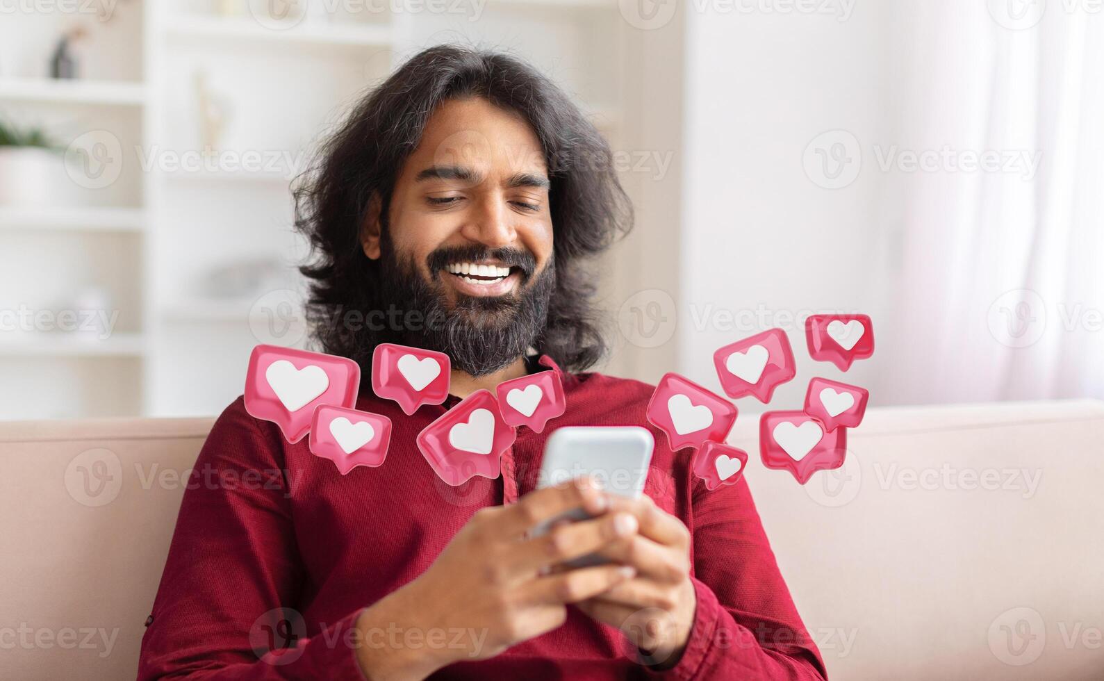 Indian man with long hair and a beard sits on a couch, smiling as he checks his smartphone. Around him are pink notification icons with white hearts, indicating that he has received many likes photo