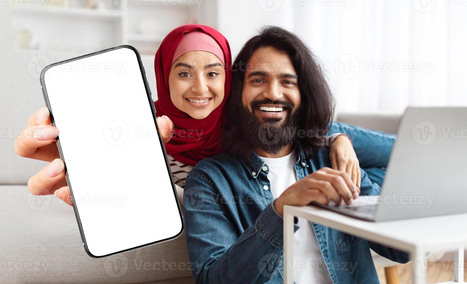 A cheerful Indian muslim couple sits closely together on a sofa, with one partner holding a phone and the other working on a laptop. They enjoy a relaxed moment at home while smiling at the camera. photo