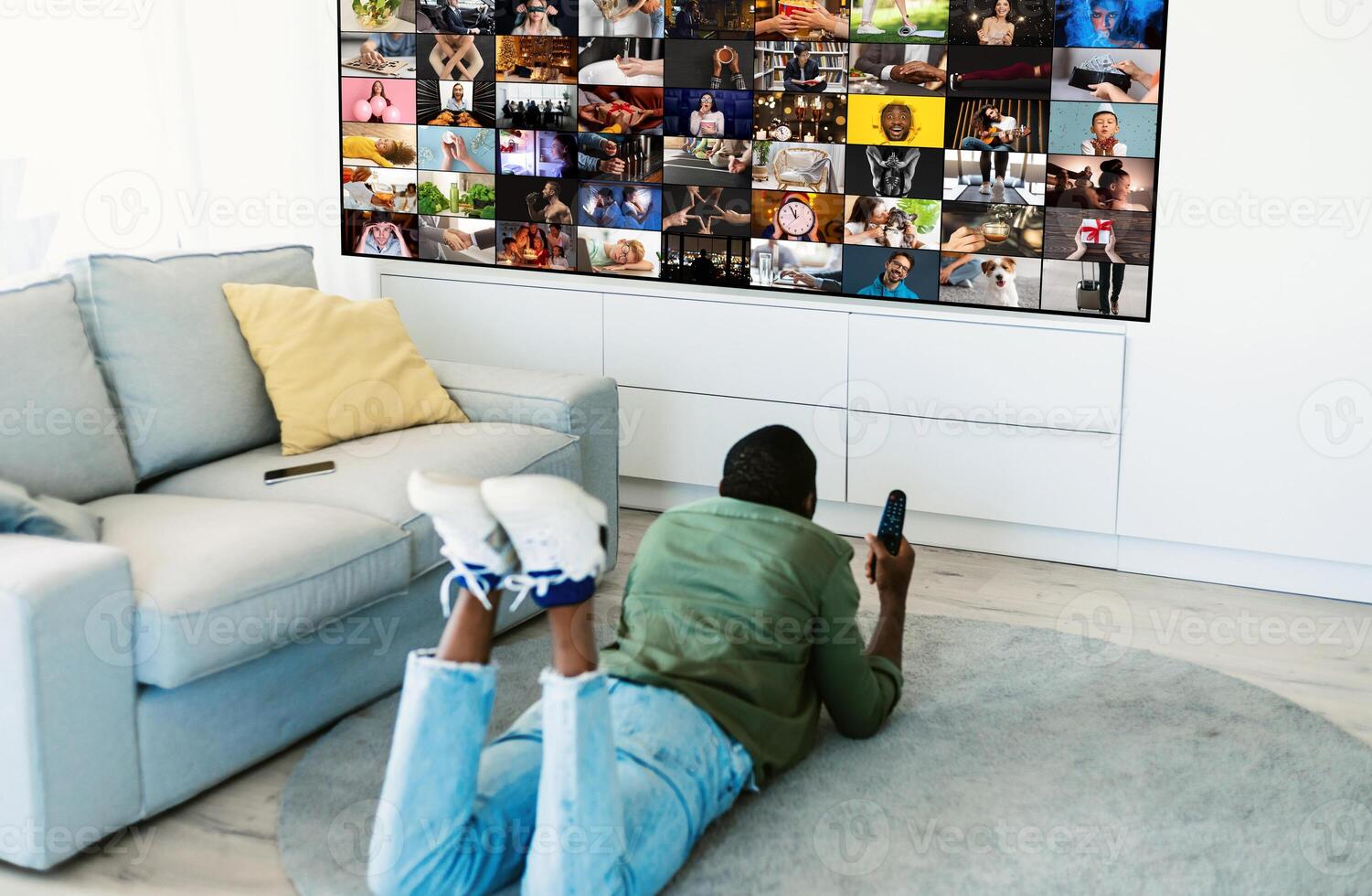 A man is lying on the floor of a living room, watching television. The television is displaying a grid of various images, and the man is holding a remote control in his hand. photo