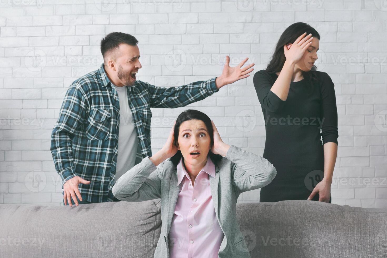 A woman sits on a couch with her hands over her ears as a man yells at another woman standing behind the couch. Woman standing behind the couch with her hand on her forehead. photo