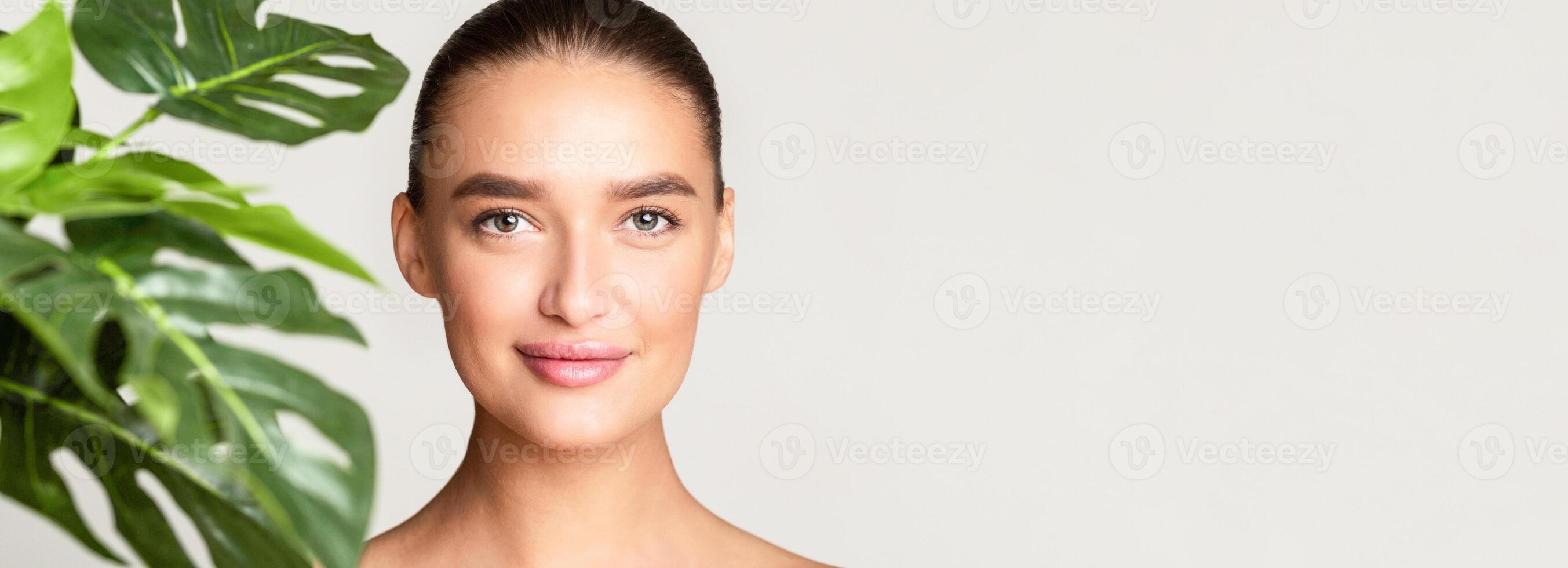 A woman face is close-up in the foreground, with a green plant in the background. She is looking towards the camera, with the plant slightly out of focus behind her, panorama, copy space photo
