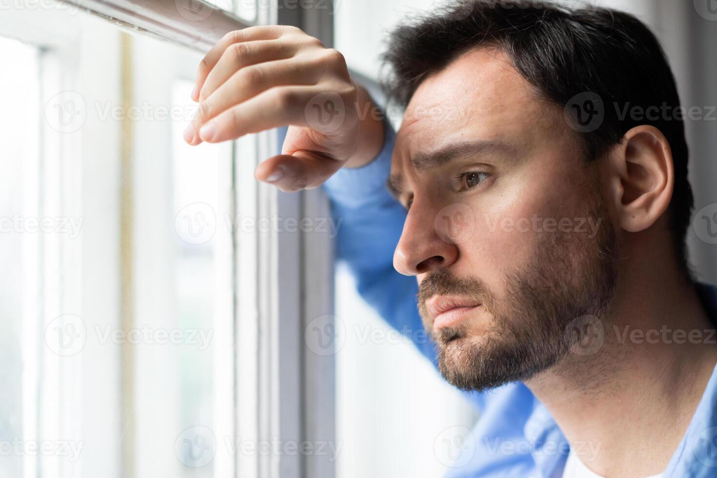 A man stands by a window, gazing out with a thoughtful expression. The natural light highlights his profile as he observes the view outside. photo