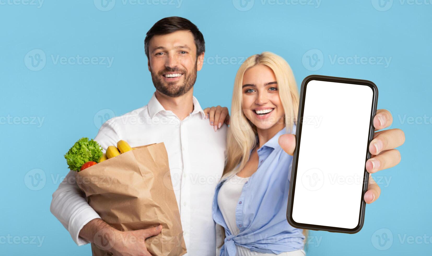 A young man and woman are standing in front of a light blue background. The man is holding a brown paper bag filled with groceries, while the woman is holding a smartphone with a blank white screen photo