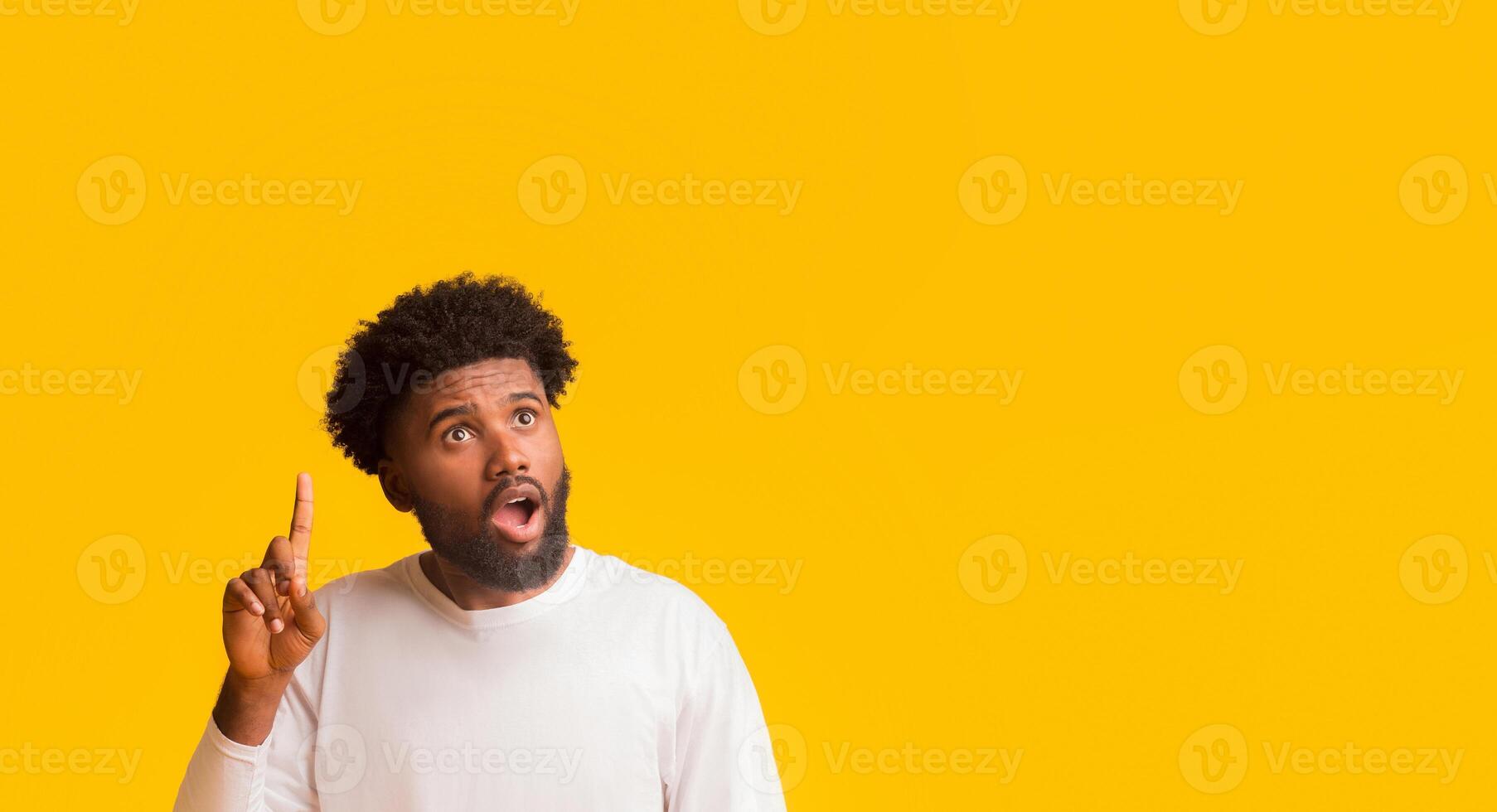 Amazed black guy with open mouth pointing finger up and looking at free space over orange background, panorama photo