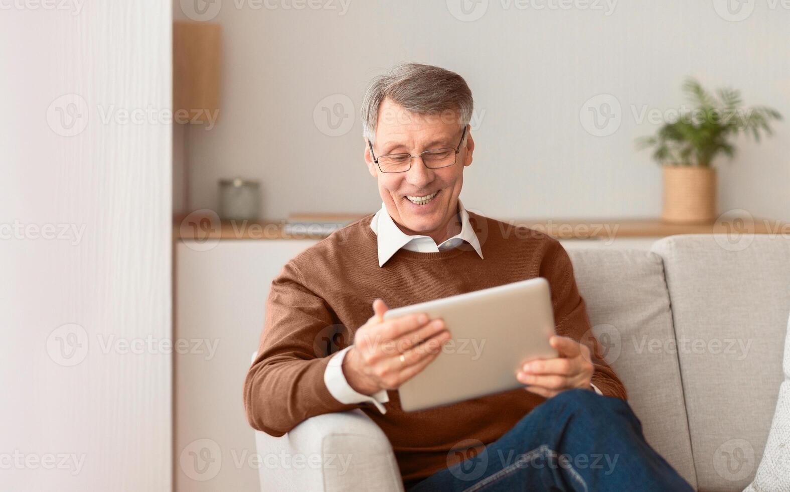 Retirement Life. Happy Mature Man Using Tablet Computer Reading Business News Sitting On Couch At Home. Selective Focus, Copy Space photo