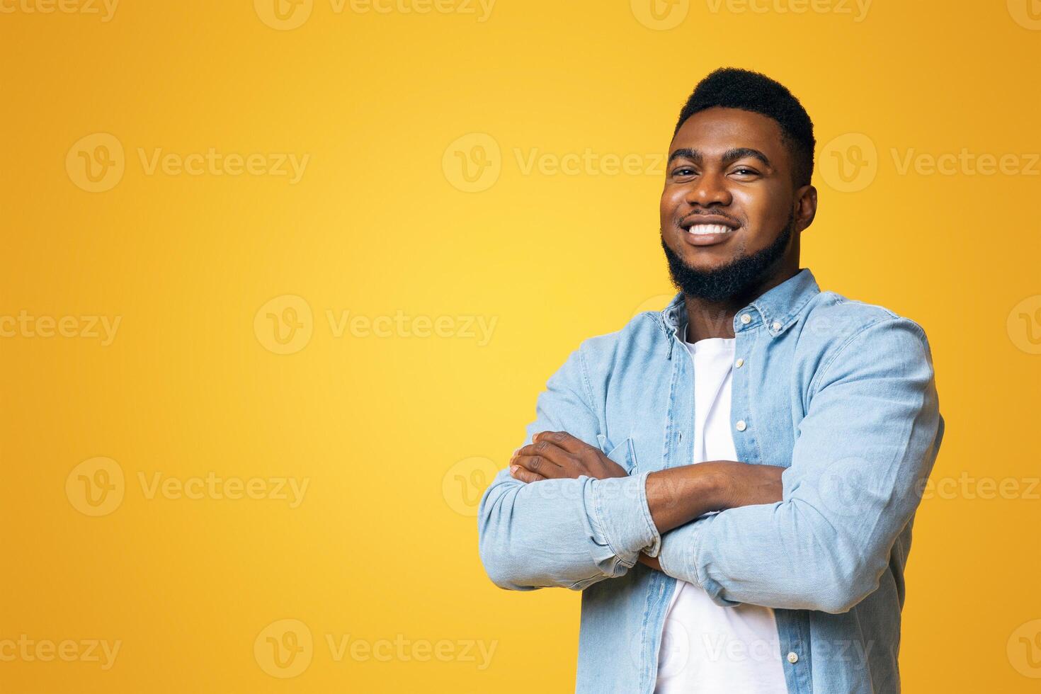 Confident black millenmial man standing with his arms crossed, smiling and looking at camera, yellow studio background with copy space photo