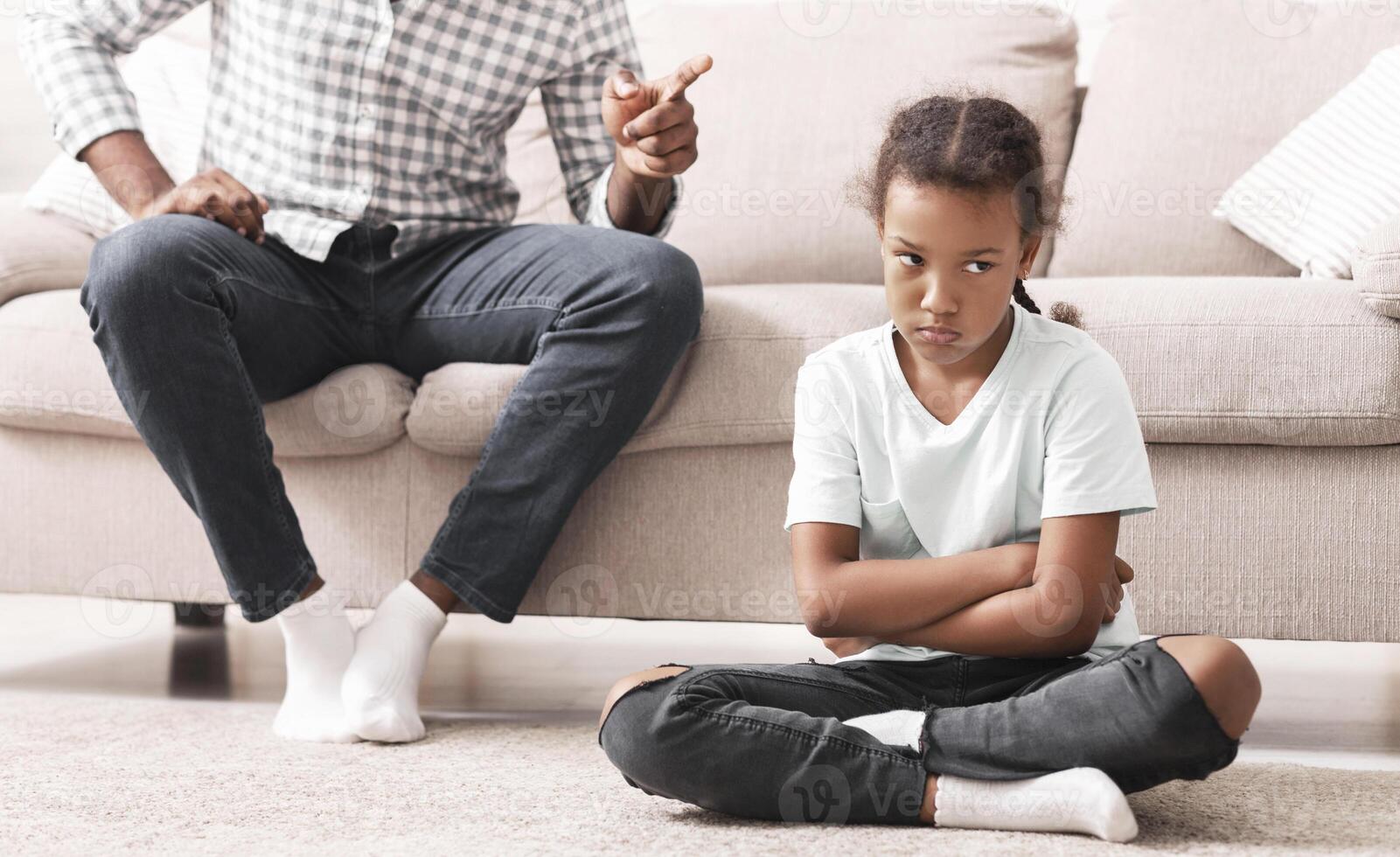 Disobedient Child. Strict black dad scolding stubborn little daughter for bad behavior at home, girl sitting on floor feeling bored and offended photo