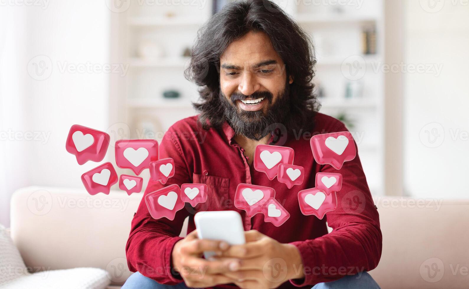 Hindu man with a long beard is sitting on a couch and smiling while looking at his smartphone. There are many heart icons surrounding him, suggesting he is receiving lots of likes photo